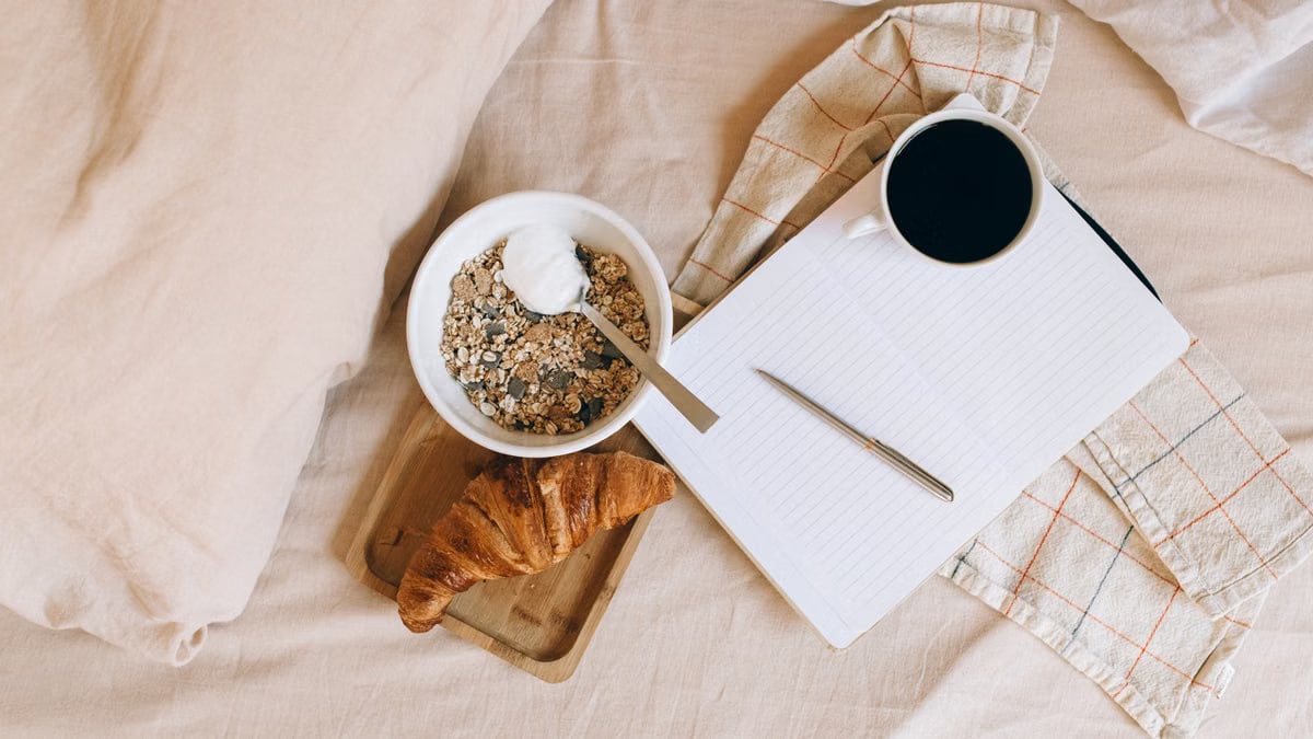 A cozy breakfast setup on a bed featuring a croissant, yogurt bowl, coffee, and notebook.